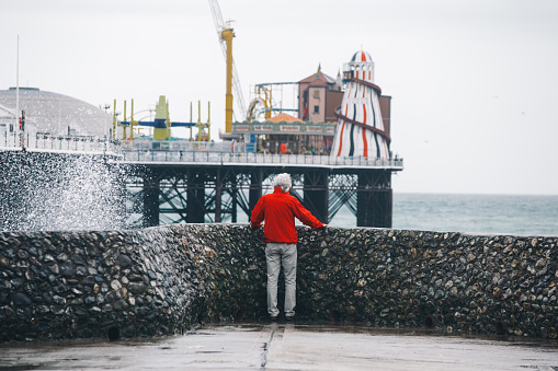 Rear view color image depicting a senior man, wearing a red jacket, standing on the end of a pier on the coastline. He is looking out to sea, and the violent waves are battering the pier so that he is surrounded by sea spray.