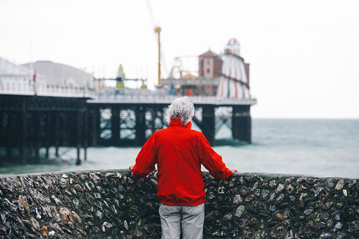 Rear view color image depicting a senior man, wearing a red jacket, standing on the end of a pier on the coastline. He is looking out to sea, as if he is deeply contemplating life.