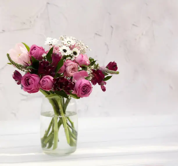 Photo of A bouquet of pink, burgundy and white flowers - alstroemeria tulips, roses, carnations, chrysanthemums in a bouquet against the background of a white cement wall of a wooden floor in spots of light.