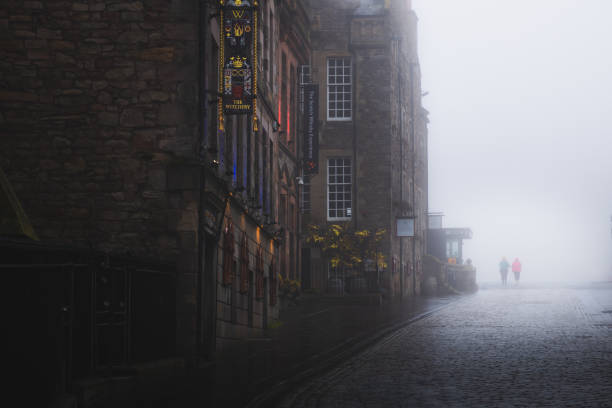 Two silhouettes emerge in moody atmospheric old town Edinburgh along the cobblestone Royal Mile in misty fog.