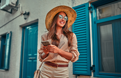 Attractive girl tourist in a blouse, sunglasses and a straw hat uses a smartphone on a background of a house with blue shutters. The concept of tourism, travel, leisure.