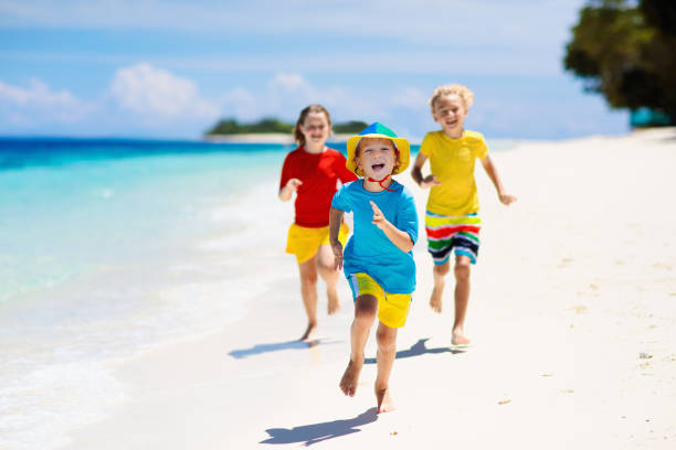 Kids playing on tropical beach. Children swim and play at sea on summer family vacation. Sand and water fun, sun protection. Little child running and jumping at ocean shore.