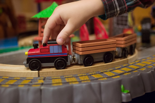 Children wooden playthings. Close up side photo of childs hand controlling toy train on the way. Little boy playing with modern wooden railway. rail road and locomotive in the play room with many toys