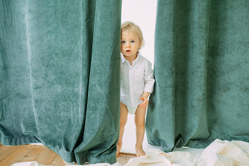 Toddler boy hides behind a curtain in a room and looks out from behind it.