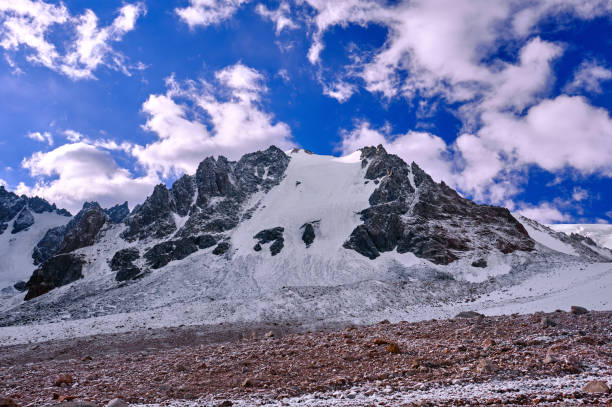 Rocky peak with a powerful glacier towering above the snow-covered moraine in the highlands on the background of a blue sky with clouds