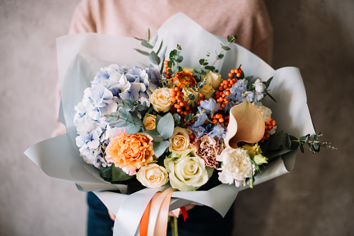Very nice young woman holding big beautiful blossoming bouquet of fresh hydrangea, roses, carnations, calla lilies, berries, eucalyptus, flowers in blue pink, cream and orange colors