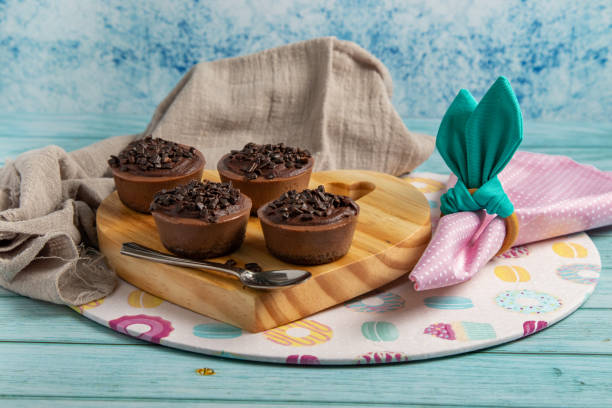 Four small chocolate-flavored brigadier cakes on an Easter table with pink napkin, napkin ring similar to rabbit ears, a themed sousplat and wooden stand. Blue background.