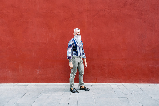 Portrait of handsome well dressed Caucasian senior man with gray beard and hair with red wall behind him.