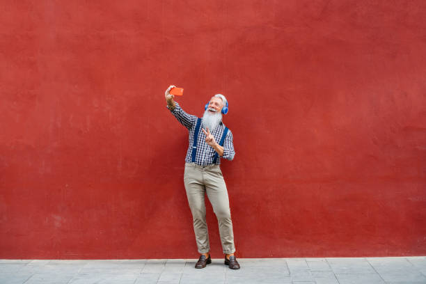 Handsome senior man making selfie Handsome well dressed senior man with gray beard and hair making selfie while leaning on red wall. man beard plaid shirt stock pictures, royalty-free photos & images