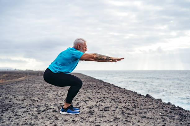Handsome senior man doing squats by the sea Handsome muscular Caucasian senior man doing squats by the sea. DisruptAgingCollection stock pictures, royalty-free photos & images