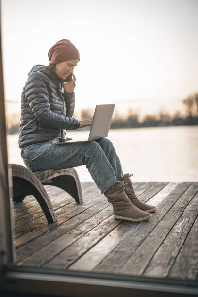 istock Woman in jacket standing on balcony while working on laptop and talking on phone 1309757440