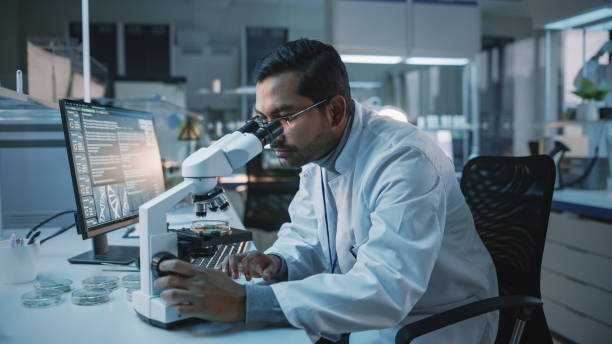 istock Medical Research Scientist Conducts DNA Experiments Under a Microscope and Writes Results on Desktop Computer in a Biological Science Laboratory. Diverse Multiethnic Lab with Engineers in White Coats. 1309776176