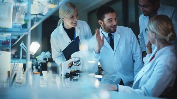 istock Surprized Female Reseach Scientist Makes an Important Discovery While Researching Samples Under the Microscope. Happy Colleagues Give High Five and Share Success with Fellow Bioengineers. 1309776191
