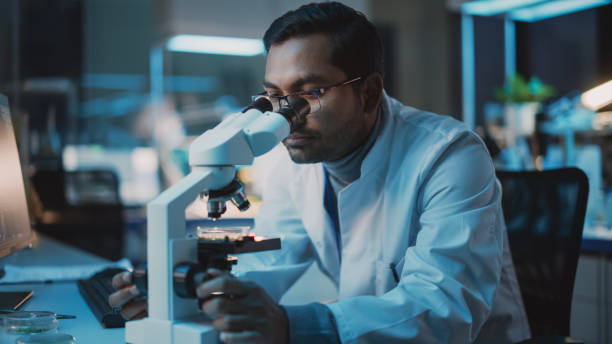 istock Medical Research Scientist Conducts DNA Experiments Under a Digital Microscope in a Biological Applied Science Laboratory. Multiethnic Lab Engineer in Glasses and White Coat Working on Vaccine. 1309776214