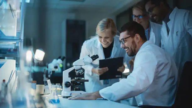 Photo of Happy Male Reseach Scientist Makes an Important Discovery While Researching Samples Under the Microscope. Happy Colleagues Congratulate it and Share Success with Fellow Bioengineers.