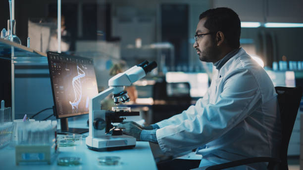 istock Medical Research Scientist is Using Desktop Computer Writes Results, Conducts DNA Experiments Under Electron Microscope in a Science Laboratory. Diverse Multiethnic Lab Engineer in White Coat. 1309776246