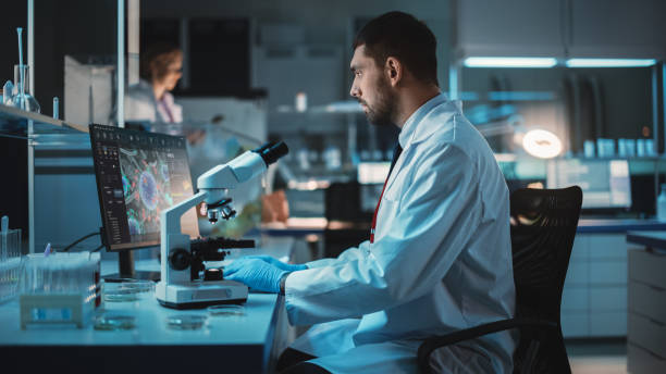 medizinischer forscher führt dna-experimente unter einem mikroskop durch und schreibt ergebnisse auf desktop-computer in einem biological science laboratory. schöne kaukasische labor-ingenieur in weißen mantel. - scientist research test tube lab coat stock-fotos und bilder