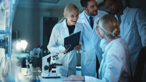 istock Happy Female Reseach Scientist Makes an Important Discovery While Researching Samples Under the Microscope. Happy Colleagues Celebrate it and Share Success with Fellow Bioengineers. 1309776277