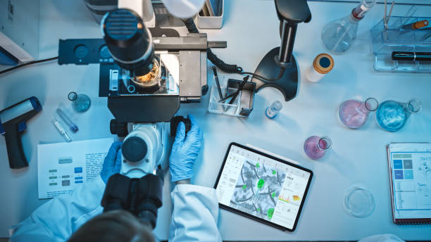 top down view of a medical research scientist in blue rubber gloves working behind table in a modern laboratory. doctor is using modern microscop for sample analysis and digital tablet with virus data - microscop imagens e fotografias de stock