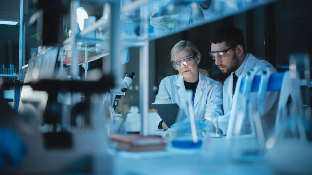 istock Female and Male Medical Research Scientists Have a Conversation While Conducting Experiments in Sample Test Tubes, Writing Analysis Results on a Tablet Computer. Modern Biological Science Laboratory. 1309776312