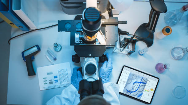 top down view of a medical research scientist in blue rubber gloves working behind table in a modern laboratory. doctor is using modern microscop for sample analysis and digital tablet to save data. - microscop imagens e fotografias de stock