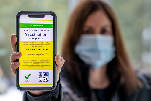 Vaccinated woman using digital vaccine passport app in mobile phone at customs for travel during Covid-19 pandemic. She is showing her immunization certificate and passport to the customs official. She is wearing a protective face mask.