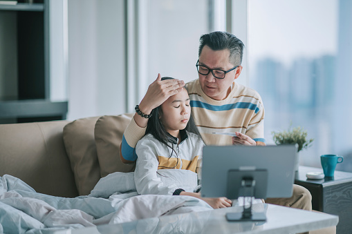 asian chinesse father taking temperature for his sick daughter on sofa living room videocall with his doctor via digital tablet