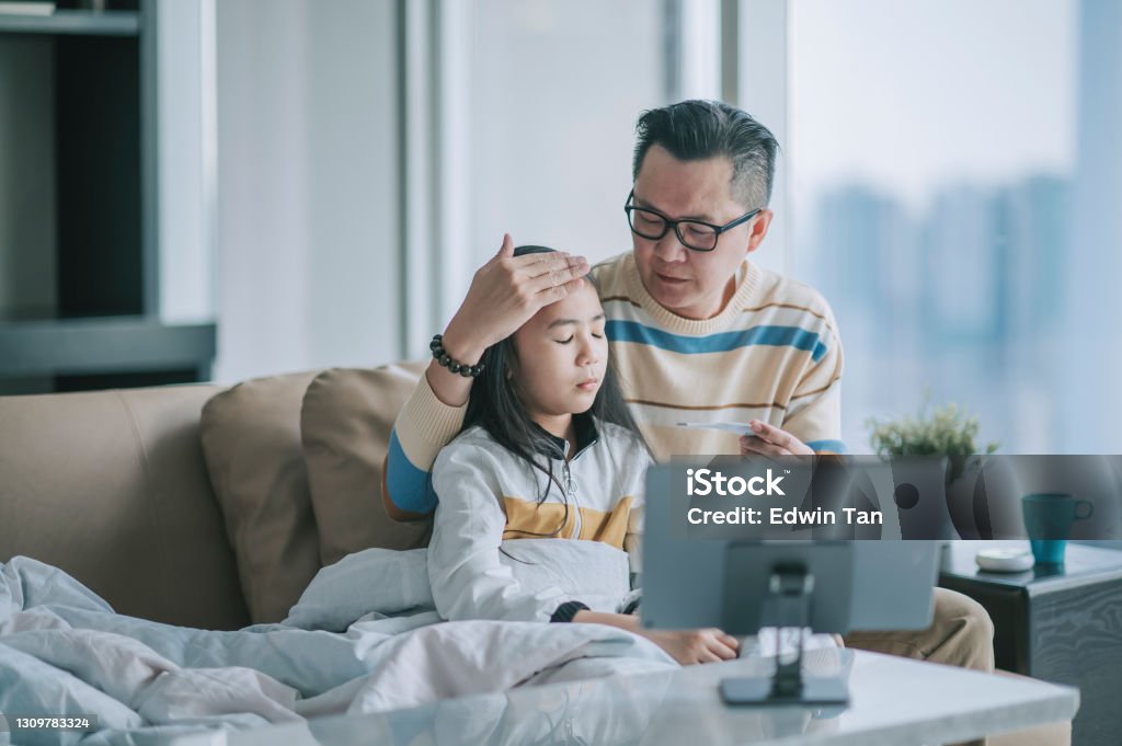 asian chinesse father taking temperature for his sick daughter on sofa living room videocall with his doctor via digital tablet Telemedicine Stock Photo