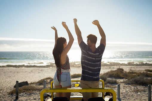 Happy caucasian couple in beach buggy by the sea with hands up. beach break on summer holiday road trip.