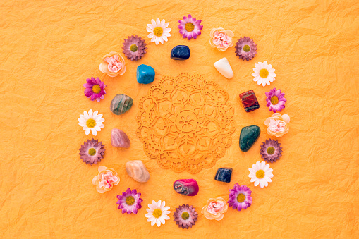 Circle of meditation or healing stones with flowers and a golden mandala in the centre on yellow crumpled tissue paper. Healing stones are: Bronzite, Aventurin, Tiger-eye, Lapis lazuli, Jade, pink Agate,Rose quartz, Turquoise, Dumortierite, dark green Aventurin. Grain added. Part of a series.
