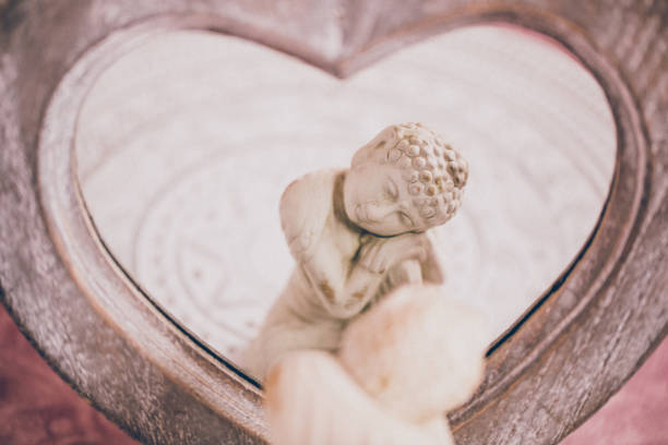 White Buddha figurine reflecting in a heart shape mirror in soft  ambience. For mindfulness and wisdom concepts. Specifically helpful for mindfulness self-compassion concepts. Soft focus on the figurine in the mirror. Added grain. Part of a series.