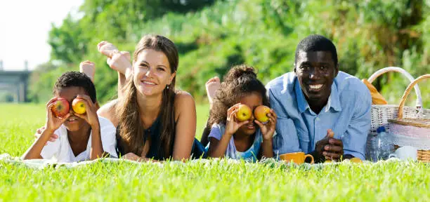 Cheerful mixed race couple with preteen children gaily spending time at picnic, lying together on green lawn