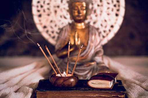 Fuming incense sticks in a candle with a very small diary on an old book in front of the part of a golden Buddha figurine. For mindfulness and wisdom concepts.  Selective focus and added grain. Part of a series.