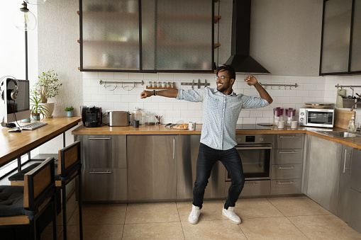 Smiling African American man dancing, having fun in modern kitchen, happy young male moving to favorite music in the morning alone, preparing breakfast, starting new day, enjoying leisure time
