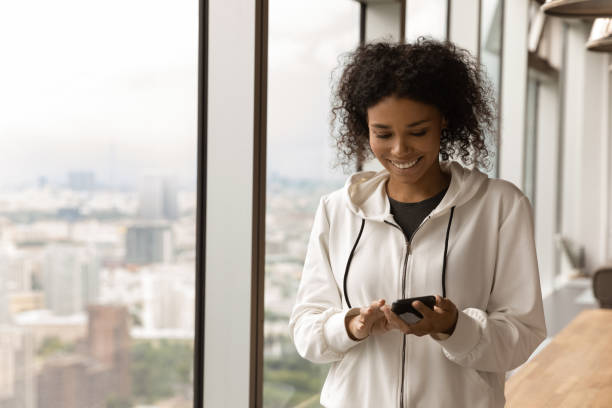 istock Smiling African American woman using phone, standing near panoramic window 1309810289