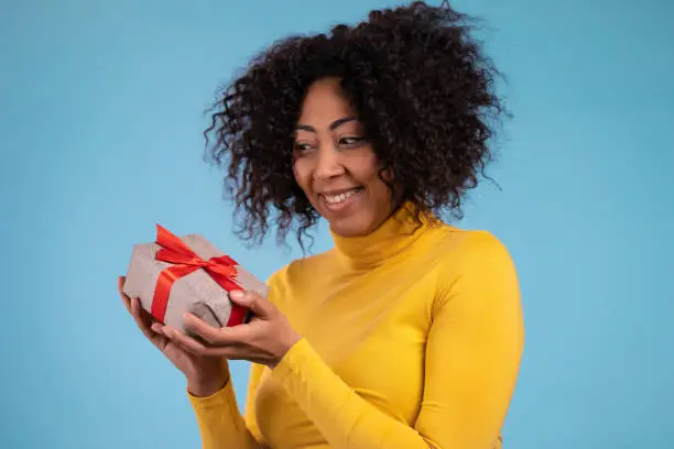 Excited african woman received gift box with bow. She is happy and flattered by attention. Girl smiling with present on blue background. Studio portrait.