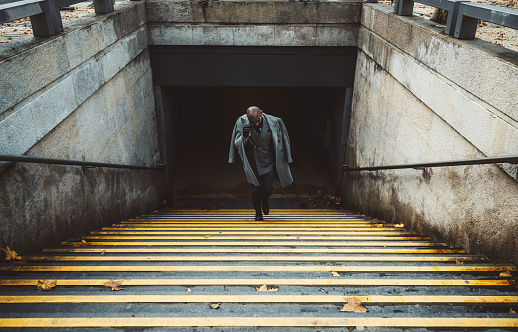 View of an elegant bald man entrepreneur climbing the stairs with yellow lines from the metro station to the street; a dapper African businessman in a coat and eyeglasses leaving an underground pass