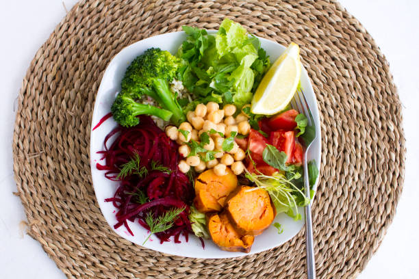 A healthy vegan buddha bowl on natural place mat.\nShot from above the bowl includes chickpeas, sweet potato, broccoli and raw and cooked vegetables.\nStock photo.