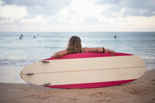 Rear view of a young adult woman sitting on the beach and leaning against her surfboard while watching the ocean waves.