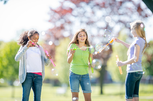 Three (3) teenage girls are outdoors at a park on a summer day. They are blowing bubbles using a bubble wand. They are dressed casually.