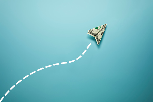 This is a photograph of United States One Dollar Bill in the shape of a paper airplane on a blue background. There is a dotted chalk line drawn in a fun path behind the plane to create a creative likable jet like trail. There is a lot ofspace for copy on the blue bakground and the path creates a nice border and frame for any text.
