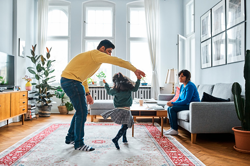 Woman looking at man and daughter while sitting on sofa. Girl is dancing with father in living room. They are spending leisure time together.