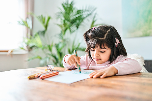 Cute girl drawing while sitting at table. She is coloring on paper. She is at home.