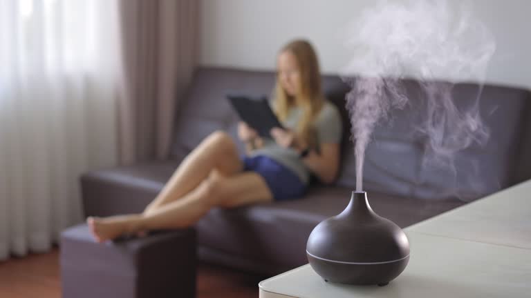 The woman in the background is sitting on a couch and a working aromatherapy diffuser is on a table in a foreground