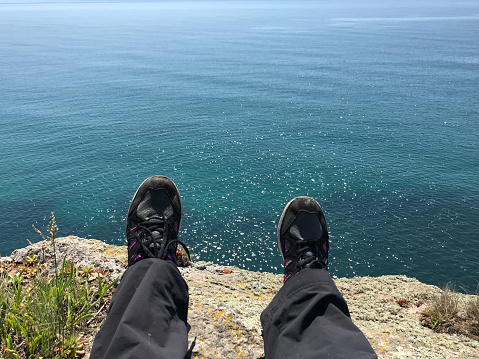 Woman's legs and feet clad in sneakers on the cliff edge above blue sea