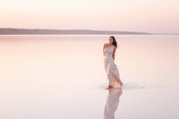 Young blonde woman in an evening airy pastel pink, powdery dress stands barefoot on white crystallized salt. Girl with natural make-up, hair is developing. Salt mining trip, walking on water at sunset joven rubia en una noche de color rosa pastel, vestido polvoriento se encuentra descalzo sobre sal cristalizada blanca. chica con maquillaje natural, el cabello se está desarrollando. viaje de minería de sal, caminando sobre el agua al atardecer