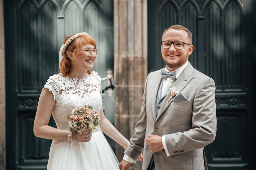 Bride and groom standing outside the church. The redheaded bride is holding a flower bouquet. Both are smiling happily.