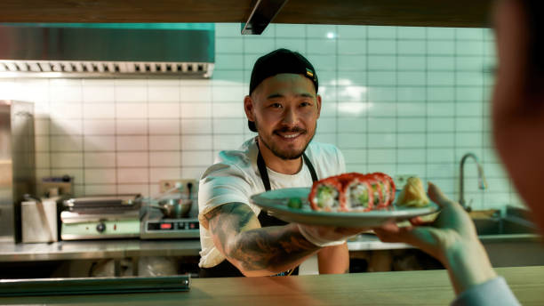 веселый молодой шеф-повар японского ресторана улыбается, давая суши-роллы официантке, работающей на кухне - restaurant dinner waitress dining стоковые фото и изображения