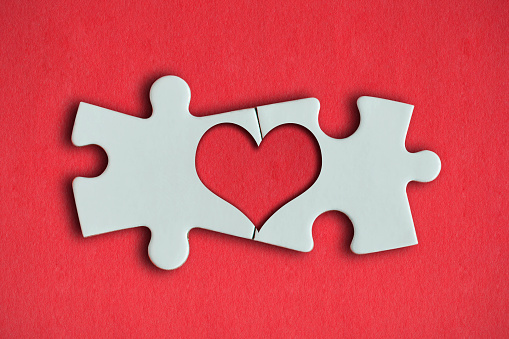 Two white puzzle pieces comes together to create a heart shape on the red background. Horizontal composition with enough copy space for your text.