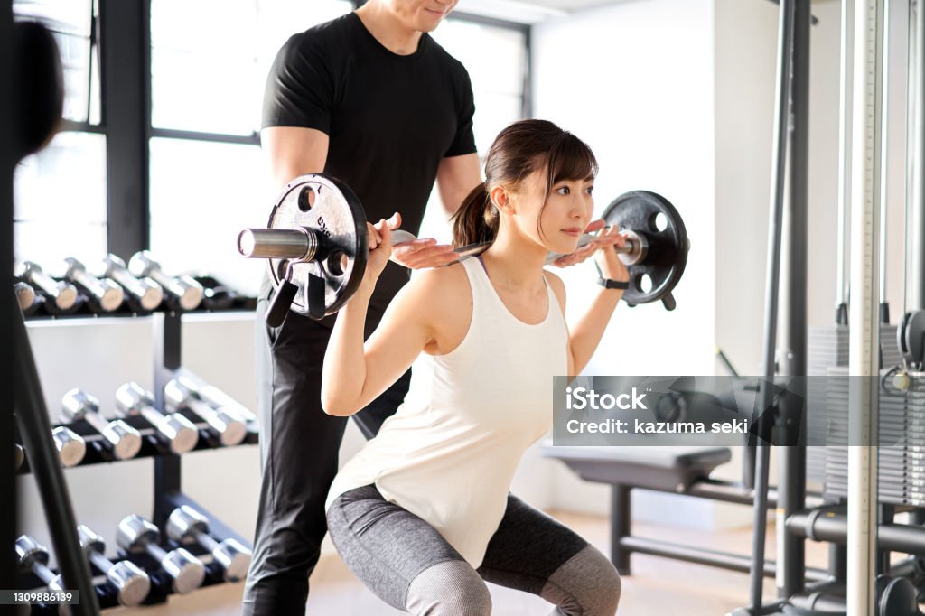 Asian woman doing barbell squats with the assistance of a trainer Gym Stock Photo